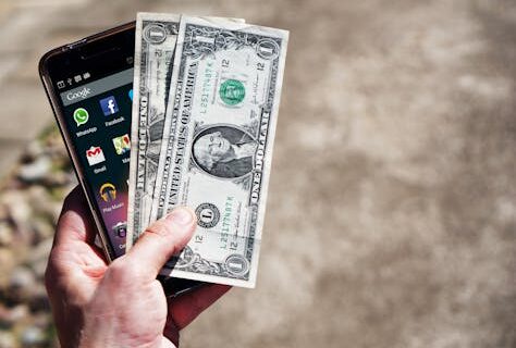 Close-up of a hand holding US dollar bills and a smartphone outdoors, showcasing financial technology.